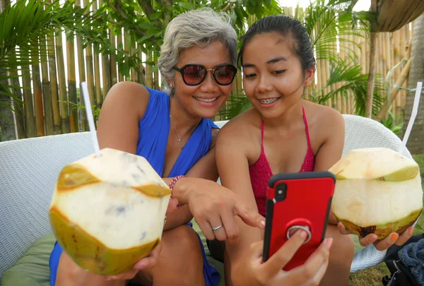 Feliz y hermosa asiática indonesia adolescente tomando selfie con teléfono móvil disfrutar de vacaciones beber coco en tropical resort jardín junto con su madre —  Fotos de Stock