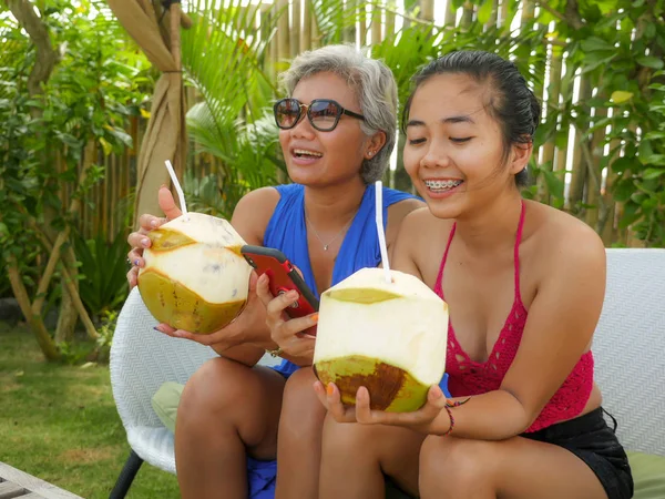 Feliz y hermosa asiática indonesia adolescente chica usando móvil disfrutando de vacaciones de verano beber coco en tropical resort jardín junto con su madre —  Fotos de Stock