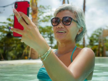 mutlu ve çekici Asya Endonezya orta yaşlı 40'lı veya 50s kadın tropikal tatil yüzme havuzunda tatil zevk cep telefonu ile selfie fotoğraf çekmek