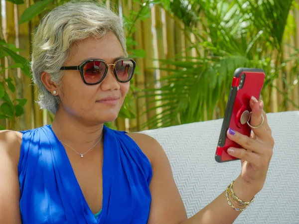 Feliz y atractivo asiático indonesia mujer relajado en tropical playa resort jardín tomando selfie en móvil teléfono sonriendo disfrutando vacaciones viaje — Foto de Stock