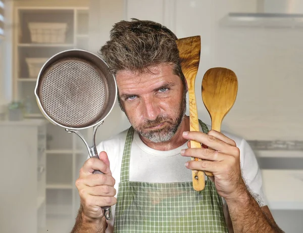 attractive unhappy and overwhelmed home cook man in apron holding spoon and pan feeling upset and lazy at house kitchen in domestic work and cooking duty