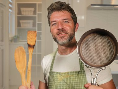Genç, mutlu ve çekici ev aşçısı önlüklü neşeli ve büyüleyici bir adam elinde tepsi ve kaşıkla modern mutfakta ev işi ve yemek pişirme hobisinin keyfini çıkarıyor.