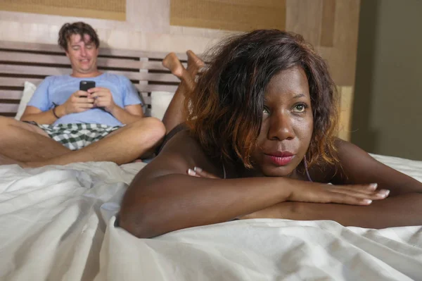 Проблема отношений в смешанной этнической паре в постели с белым мужчиной, использующим мобильный телефон в качестве интернет-наркомана, и черной афро-американской жене или подруге, испытывающей подозрения — стоковое фото