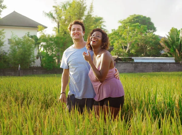 Счастливая и красивая смешанная этническая пара с черной афро-американской женщиной и привлекательным кавказским мужчиной обнимается и веселится наслаждаясь праздниками на рисовом поле — стоковое фото
