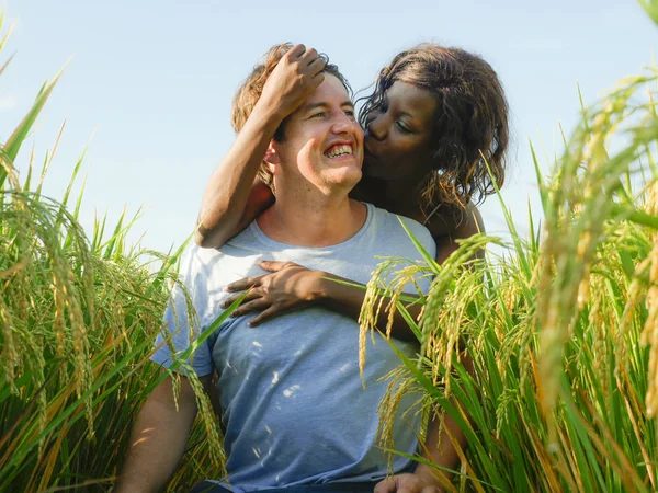 Молодая счастливая и красивая смешанная этническая пара с черной афро-американской женщиной и привлекательным кавказским мужчиной обнимается и веселится наслаждаясь праздниками на рисовом поле — стоковое фото