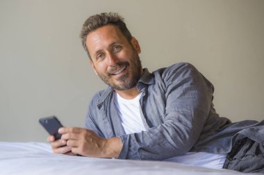 Genç, yakışıklı ve mutlu bir adam yatakta internet telefonu kullanıyor neşeli ve tatmin olmuş bir şekilde gülümsüyor internetten rahatça flört ediyor ya da sosyal medya uygulamasından zevk alıyor.