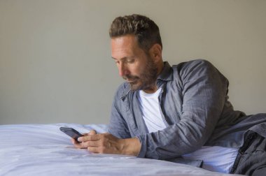 Genç, yakışıklı ve mutlu bir adam yatakta internet telefonu kullanıyor neşeli ve tatmin olmuş bir şekilde gülümsüyor internetten rahatça flört ediyor ya da sosyal medya uygulamasından zevk alıyor.