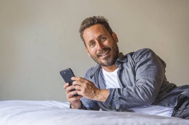 Genç, yakışıklı ve mutlu bir adam yatakta internet telefonu kullanıyor neşeli ve tatmin olmuş bir şekilde gülümsüyor internetten rahatça flört ediyor ya da sosyal medya uygulamasından zevk alıyor.