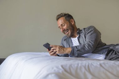 Genç, yakışıklı ve mutlu bir adam yatakta internet telefonu kullanıyor neşeli ve tatmin olmuş bir şekilde gülümsüyor internetten rahatça flört ediyor ya da sosyal medya uygulamasından zevk alıyor.