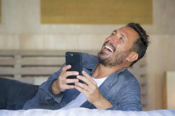 Jeune homme beau et heureux à la maison en utilisant un téléphone mobile Internet sur le lit souriant gai et satisfait se sentir détendu datant en ligne ou profiter de l'application de médias sociaux — Photo