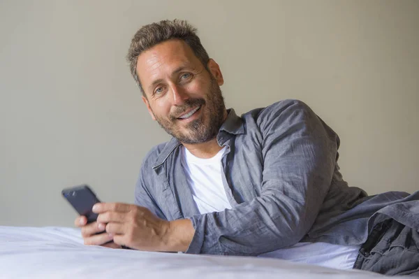 Joven guapo y feliz hombre en casa utilizando el teléfono móvil de Internet en la cama sonriendo alegre y satisfecho sensación relajado citas en línea o disfrutar de las redes sociales aplicación — Foto de Stock