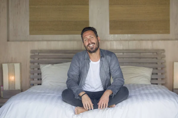 Retrato interior de 30 a 40 años hombre feliz y guapo en casa en camisa casual y jeans sentados en la cama relajado en casa sonriendo confiado y feliz sentirse positivo — Foto de Stock
