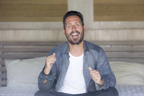 Retrato interior de 30 a 40 años hombre feliz y guapo en casa en camisa casual y jeans sentados en la cama relajado en casa sonriendo confiado y feliz sentirse positivo — Foto de Stock