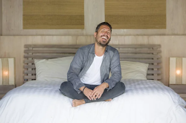 Retrato interior de 30 a 40 años hombre feliz y guapo en casa en camisa casual y jeans sentados en la cama relajado en casa sonriendo confiado y feliz sentirse positivo — Foto de Stock