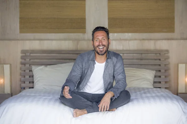 Retrato interior de 30 a 40 años hombre feliz y guapo en casa en camisa casual y jeans sentados en la cama relajado en casa sonriendo confiado y feliz sentirse positivo — Foto de Stock