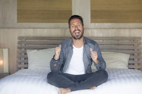 Retrato interior de 30 a 40 años hombre feliz y guapo en casa en camisa casual y jeans sentados en la cama relajado en casa sonriendo confiado y feliz sentirse positivo — Foto de Stock