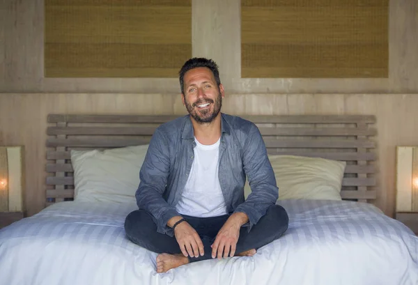 Retrato interior de los años 30 feliz y guapo hombre en casa en camisa casual y jeans sentados en la cama relajado en casa sonriendo confiado y feliz sentirse positivo — Foto de Stock