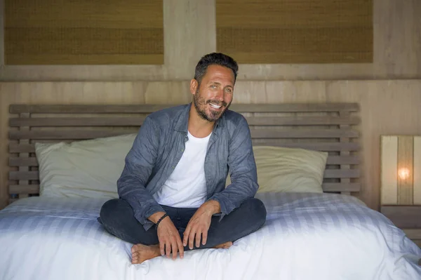 interior portrait of 30s happy and handsome man at home in casual shirt and jeans sitting on bed relaxed at home smiling confident and happy feeling positive