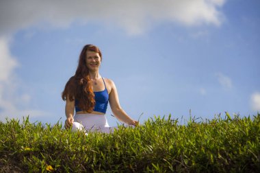 young happy and beautiful red hair woman sitting on green field relaxed on grass practicing yoga exercise outdoors smiling in wellness meditation and healthy lifestyle