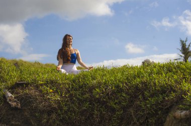 young happy and beautiful red hair woman sitting on green field relaxed on grass practicing yoga exercise outdoors smiling in wellness meditation and healthy lifestyle