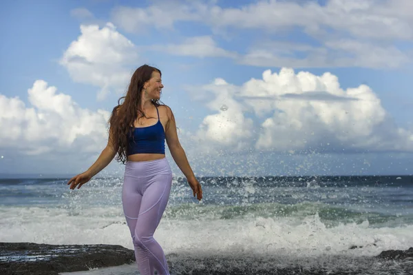 Junge glückliche und attraktive Frau mit roten Haaren, die aufgeregt Arme ausbreitet und sich frei und entspannt fühlt, nass wird, wenn die Meereswellen an ihrem Strand plantschen — Stockfoto