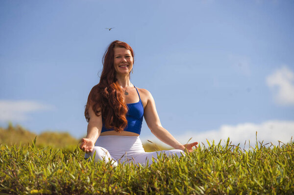 young happy and beautiful red hair woman sitting on green field relaxed on grass practicing yoga exercise outdoors smiling in wellness meditation and healthy lifestyle