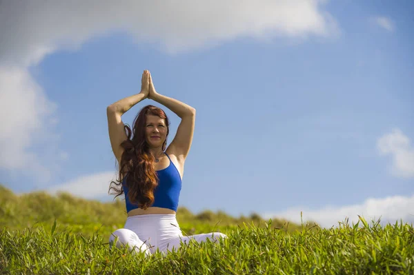 Young happy and beautiful red hair woman sitting on green field relaxed on grass practicing yoga exercise outdoors smiling in wellness meditation and healthy lifestyle — Stock Photo, Image