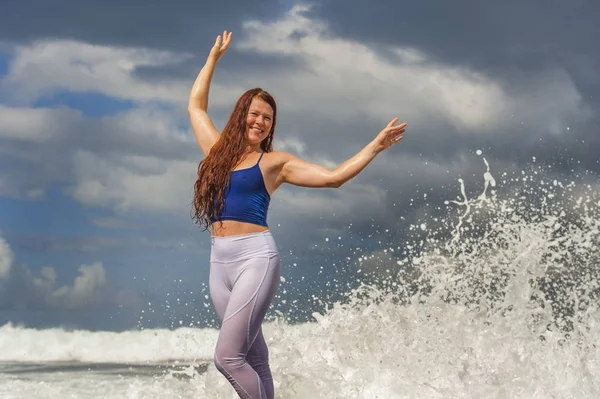 Joven feliz y atractiva mujer pelirroja jugando brazos extendidos emocionados sintiéndose libre y relajado mojándose por las olas del mar salpicando en su playa disfrutando — Foto de Stock