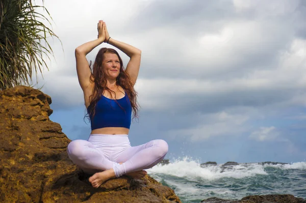 Естественный портрет молодой привлекательной и счастливой женщины с рыжими волосами, практикующей медитацию и релаксацию йоги лотоса, сидящей на берегу скалы на морском фоне — стоковое фото