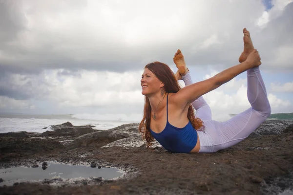 Junge glückliche attraktive und schöne Frau mit rotem Haar praktiziert Yoga-Meditation und Beweglichkeitsübungen am Strand auf Felsen vor wunderschönem Meereshintergrund — Stockfoto