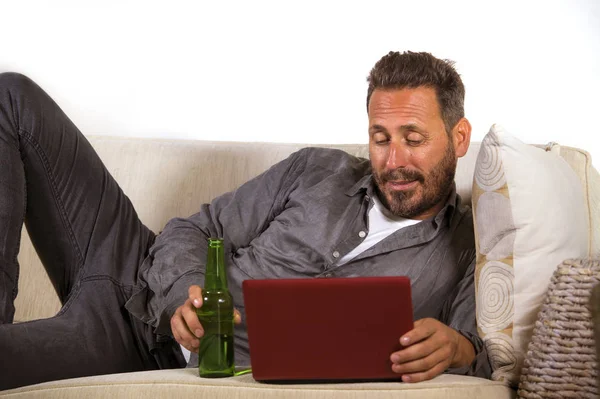 Giovane uomo indipendente bello e di successo che lavora a casa con computer portatile sdraiato rilassato sul divano vivente in rete allegra bottiglia di birra da bere — Foto Stock