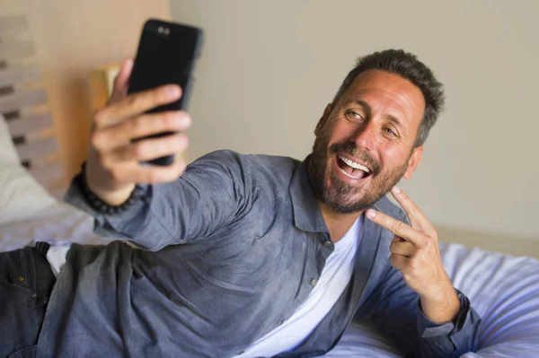 Portrait de style de vie de jeune homme attrayant et heureux des années 30 ou 40 prenant des photos selfie avec téléphone portable souriant geste ludique dans la chambre à coucher à la maison couché sur le lit — Photo