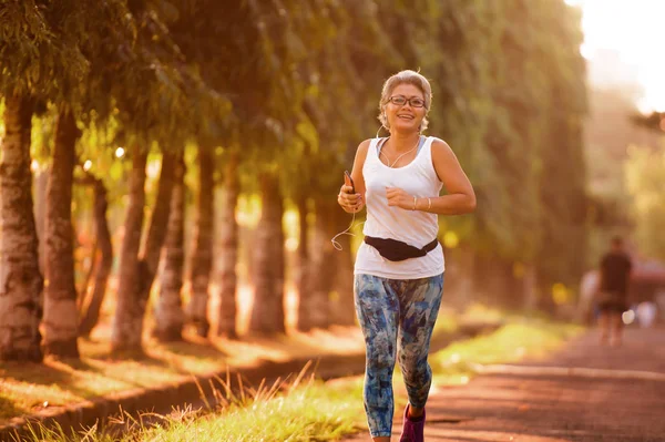 Idade média 40 ou 50 anos mulher feliz e atraente com formação de cabelos grisalhos no parque da cidade com árvores verdes ao nascer do sol fazendo corrida e jogging treino em cuidados de saúde — Fotografia de Stock