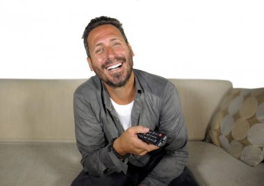 Genç, çekici ve mutlu bir adam evinde gülüyor televizyon programı ya da komedi dizisi seyrediyor oturma odasındaki koltukta rahat ve neşeli oturuyor.