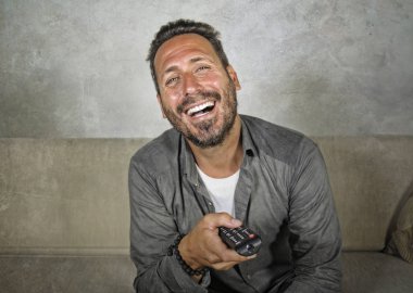 Genç, çekici ve mutlu bir adam evinde gülüyor televizyon programı ya da komedi dizisi seyrediyor oturma odasındaki koltukta rahat ve neşeli oturuyor.