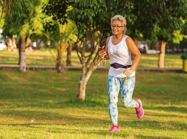 Orta yaşlı 40'lı veya 50'li yaşlarda mutlu ve çekici kadın güneş doğarken yeşil ağaçlar ile şehir parkında gri saç eğitimi ile çalışan ve sağlık koşu egzersiz yapıyor — Stok fotoğraf