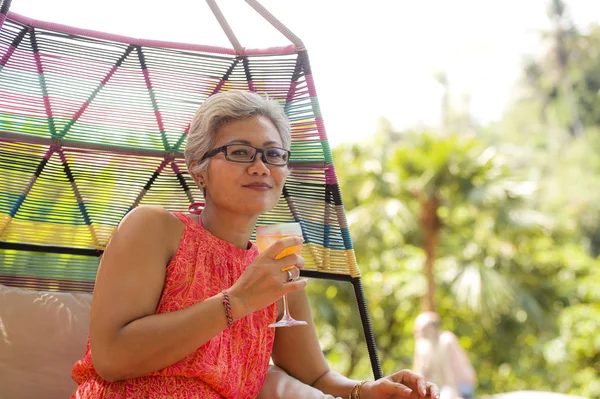 Estilo de vida natural retrato de la atractiva y feliz de los años 40 o 50 de mediana edad mujer asiática con el pelo gris y elegante vestido de verano disfrutando de la bebida en la piscina resort hamaca relajado — Foto de Stock