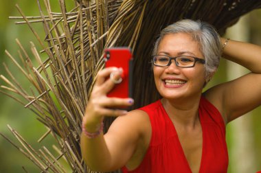 güzel tropikal ormanda cep telefonu ile selfie fotoğraf çekmek gri saçlı ve şık elbise ile çekici ve mutlu orta yaşlı 40'lı veya 50'li Asyalı kadın