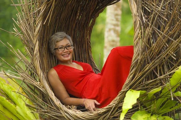 Atraente e feliz idade média 40 ou 50 anos Asiático mulher turística com cabelos grisalhos e elegante vestido vermelho sentado ao ar livre na selva tropical relaxado admirando beleza da natureza — Fotografia de Stock