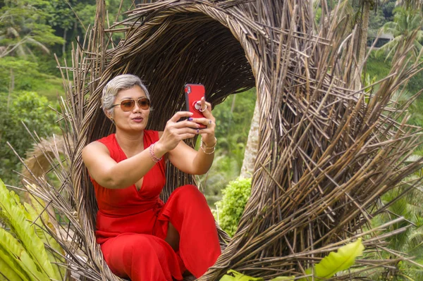 Atractiva y feliz de 40 o 50 años de edad mujer asiática con el pelo gris y vestido elegante tomar foto selfie con teléfono móvil al aire libre en la hermosa selva tropical —  Fotos de Stock