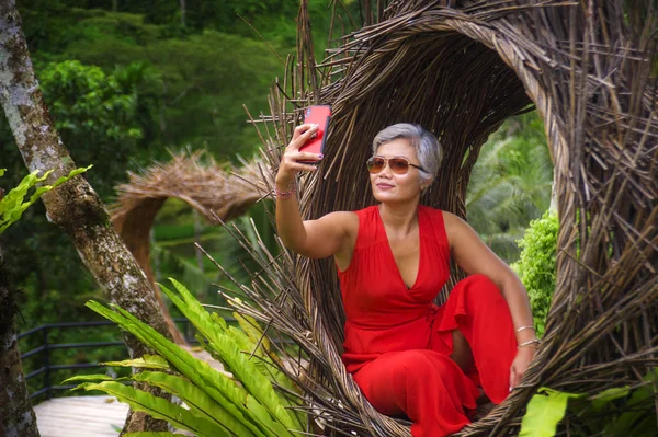 Atractiva y feliz de 40 o 50 años de edad mujer asiática con el pelo gris y vestido elegante tomar foto selfie con teléfono móvil al aire libre en la hermosa selva tropical —  Fotos de Stock