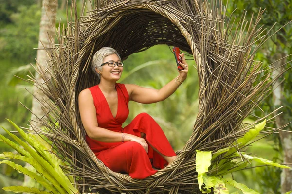 Atraente e feliz idade média 40 ou 50 anos mulher asiática com cabelos grisalhos e vestido elegante tirar foto selfie com telefone celular ao ar livre na bela selva tropical — Fotografia de Stock