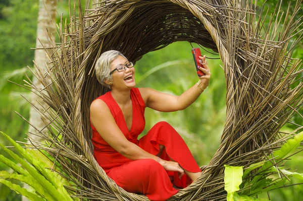 Atractiva y feliz de 40 o 50 años de edad mujer asiática con el pelo gris y vestido elegante tomar foto selfie con teléfono móvil al aire libre en la hermosa selva tropical —  Fotos de Stock