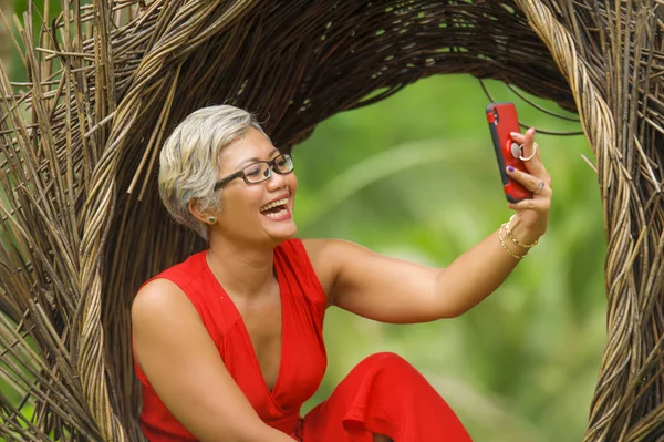Atractiva y feliz de 40 o 50 años de edad mujer asiática con el pelo gris y vestido elegante tomar foto selfie con teléfono móvil al aire libre en la hermosa selva tropical —  Fotos de Stock