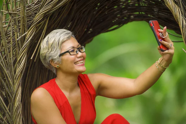 Atractiva y feliz de 40 o 50 años de edad mujer asiática con el pelo gris y vestido elegante tomar foto selfie con teléfono móvil al aire libre en la hermosa selva tropical —  Fotos de Stock