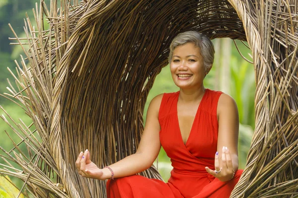 Atractiva y feliz mujer asiática de 40 o 50 años de edad en elegante y hermoso vestido rojo practicando yoga relajación y meditación en la selva tropical —  Fotos de Stock