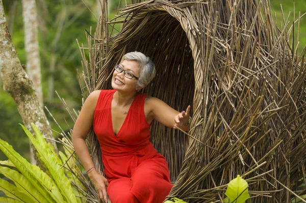 Atractivo y feliz de mediana edad de 40 o 50 años mujer turista asiática con el pelo gris y elegante vestido rojo sentado al aire libre en la selva tropical relajado admirando la belleza de la naturaleza —  Fotos de Stock