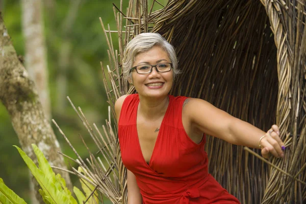 Atractivo y feliz de mediana edad de 40 o 50 años mujer turista asiática con el pelo gris y elegante vestido rojo sentado al aire libre en la selva tropical relajado admirando la belleza de la naturaleza — Foto de Stock