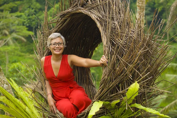 Atractivo y feliz de mediana edad de 40 o 50 años mujer turista asiática con el pelo gris y elegante vestido rojo sentado al aire libre en la selva tropical relajado admirando la belleza de la naturaleza —  Fotos de Stock