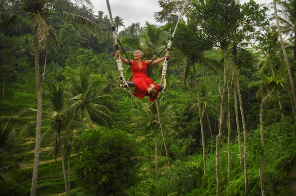 Atraente feliz meia idade 40s ou 50s asiático indonésio mulher com cabelos grisalhos equitação floresta tropical swing despreocupado balançando e desfrutar tropical selva aventura — Fotografia de Stock